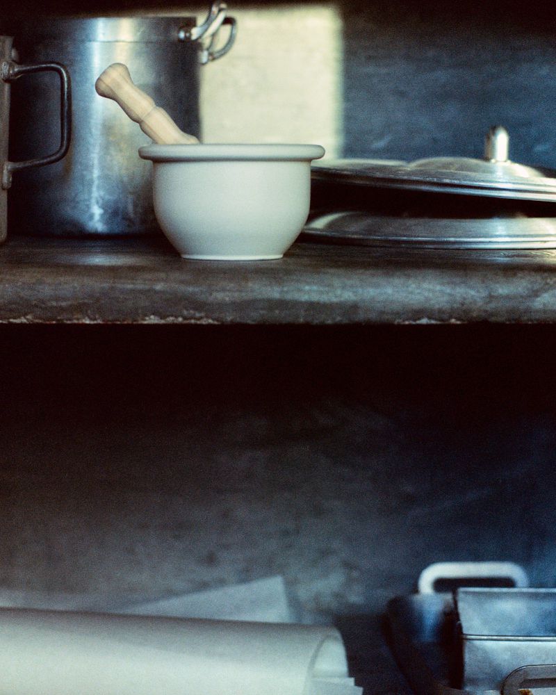 Still life shot on a rustic shelf with a white mortar and pestle, metal pots, and cookware items in a moody, vintage style.