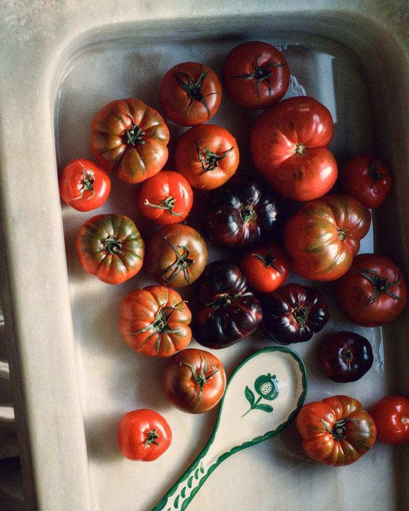 Overhead shot of an assortment of ripe heirloom tomatoes in a light-colored sink, next to a cream-colored serving spoon with green detailing.