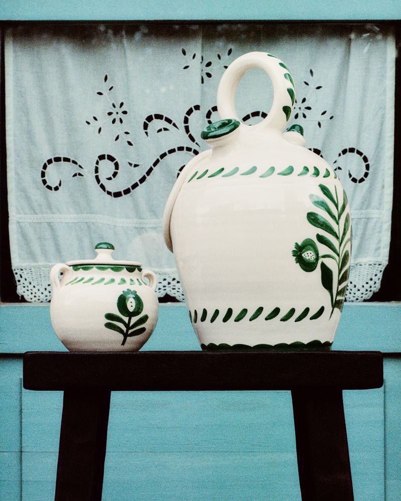 Still life of a large white ceramic pitcher and a small covered bowl with green floral decoration on a blue painted table against an embroidered curtain backdrop.