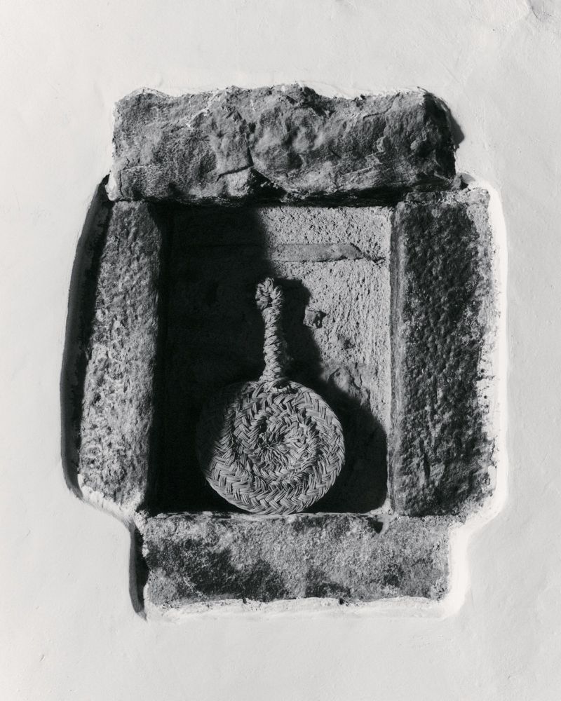 Monochrome image of a circular woven object hanging in a stone niche, creating a study in textures and light.