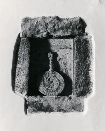 Monochrome image of a circular woven object hanging in a stone niche, creating a study in textures and light.