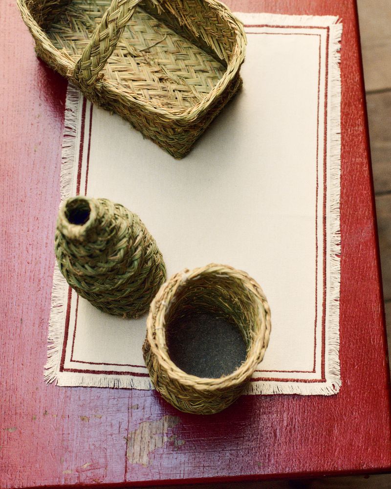Still life of woven baskets and a vase on a red wooden table with a fringed cloth, showcasing textures and craftsmanship.