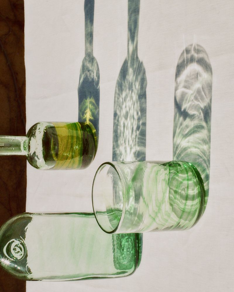Still life of translucent glassware lit in shadows with various shades of green and yellow against a white backdrop and wooden surface.