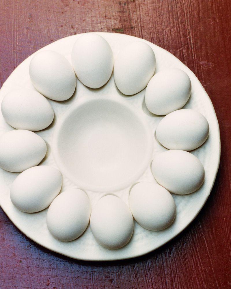Overhead shot of white eggs arranged on a white ceramic egg holder atop a dark red wooden surface, creating a minimalist food still life.