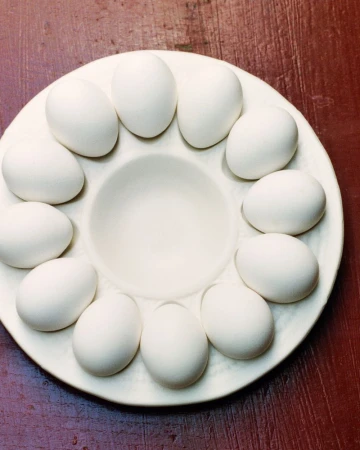 Overhead shot of white eggs arranged on a white ceramic egg holder atop a dark red wooden surface, creating a minimalist food still life.