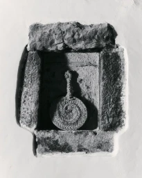 Black and white image of a woven circular pad with a looped handle nested inside a rough stone wall niche.