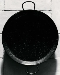 Overhead black and white photograph of a speckled black paella pan hanging against a textured tiled wall.