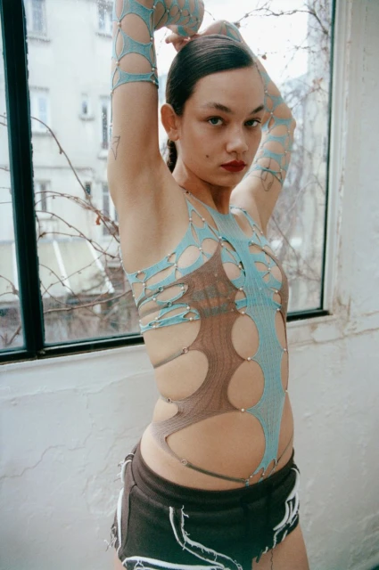Adult female in a blue cutout top and black shorts poses indoors against a window with an urban view and architectural details visible.