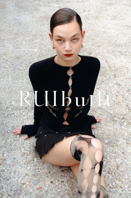 Woman in a black outfit poses on a concrete surface. The model is wearing a cardigan, skirt, patterned stockings, and jewelry.