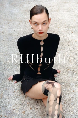 Woman in a black outfit poses on a concrete surface. The model is wearing a cardigan, skirt, patterned stockings, and jewelry.