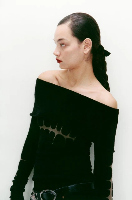 Brunette woman with red lipstick poses in off-shoulder black top with braided hair, against white backdrop.