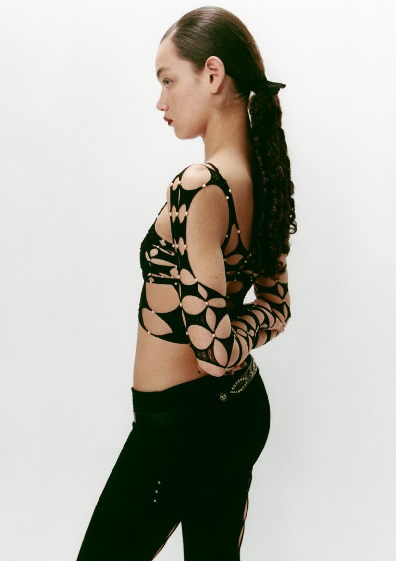 Side profile of a woman with curly ponytail wearing a patterned crop top and black pants against a plain white studio background.