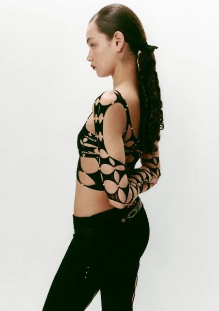 Side profile of a woman with curly ponytail wearing a patterned crop top and black pants against a plain white studio background.