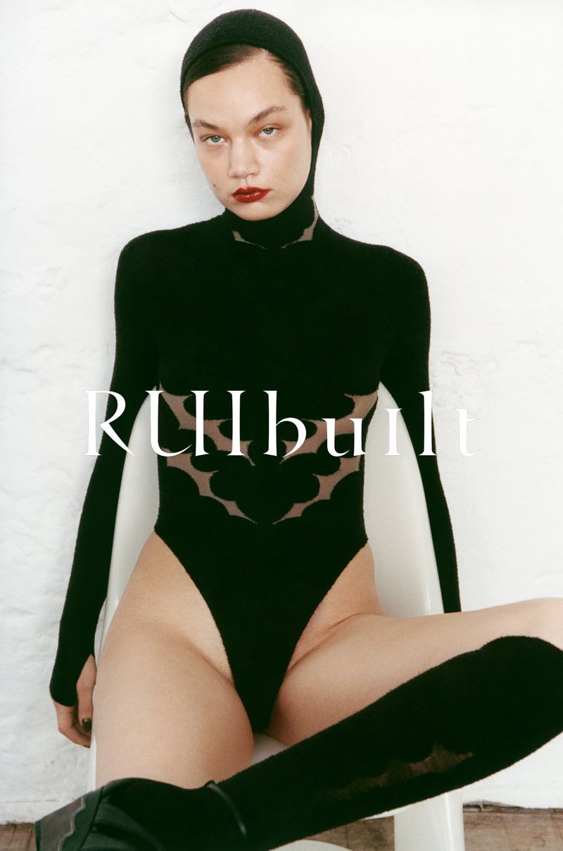 A slender woman poses in a black bodysuit and thigh-high socks against a white wall in a fashion photography studio shot.
