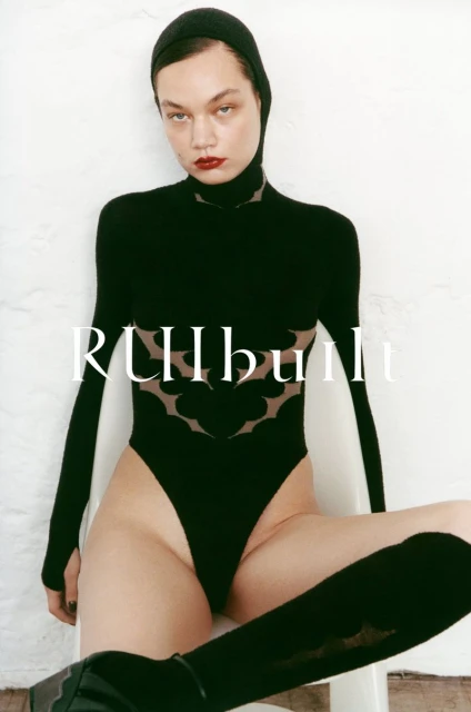 A slender woman poses in a black bodysuit and thigh-high socks against a white wall in a fashion photography studio shot.