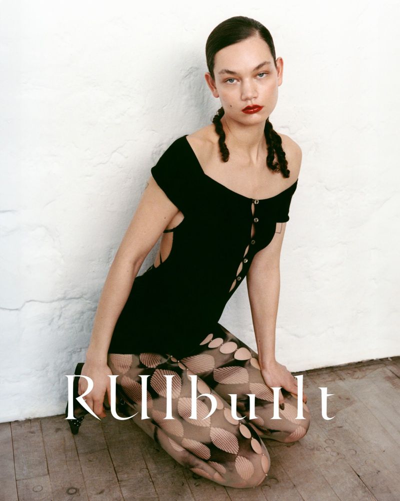 A woman in a black mini dress and patterned tights poses against a white wall in a fashion editorial-style photograph.