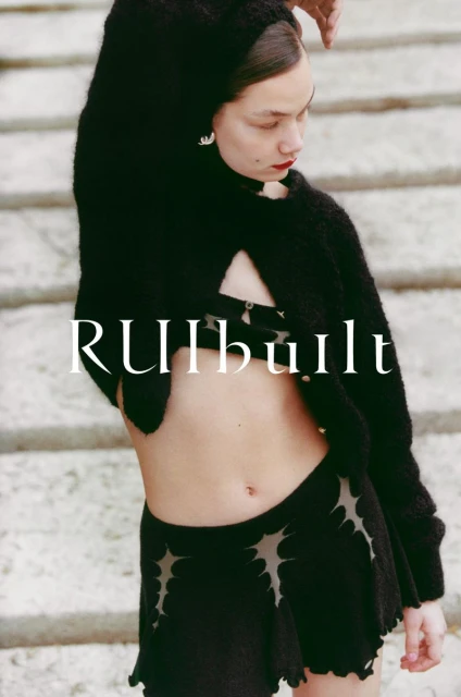 Woman in a black knit cardigan and skirt with cutout details poses against a light stone staircase, showcasing high-fashion style.