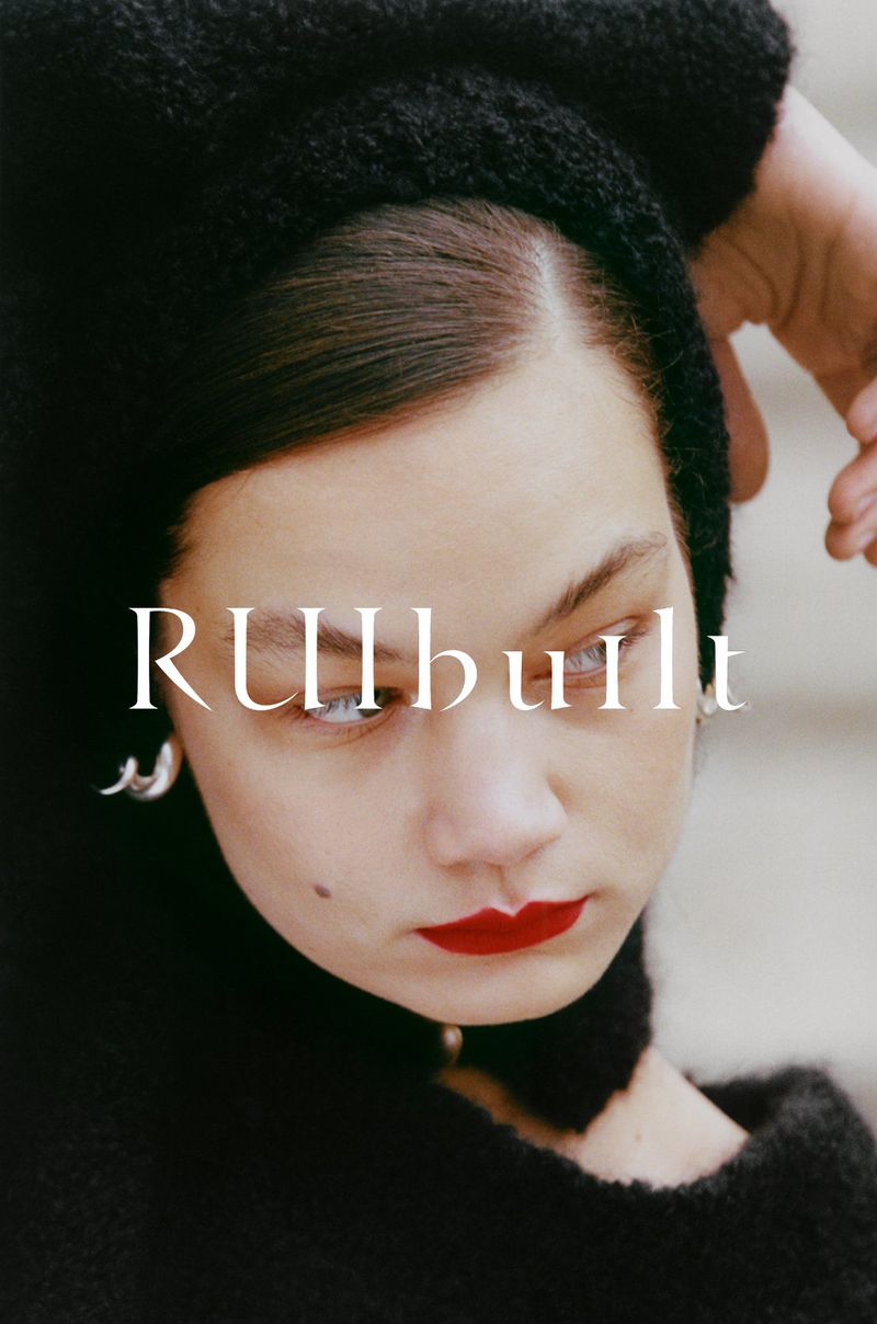 Close-up portrait of a woman with red lipstick, wearing a black textured hood or collar, accessorized with modern silver earrings.