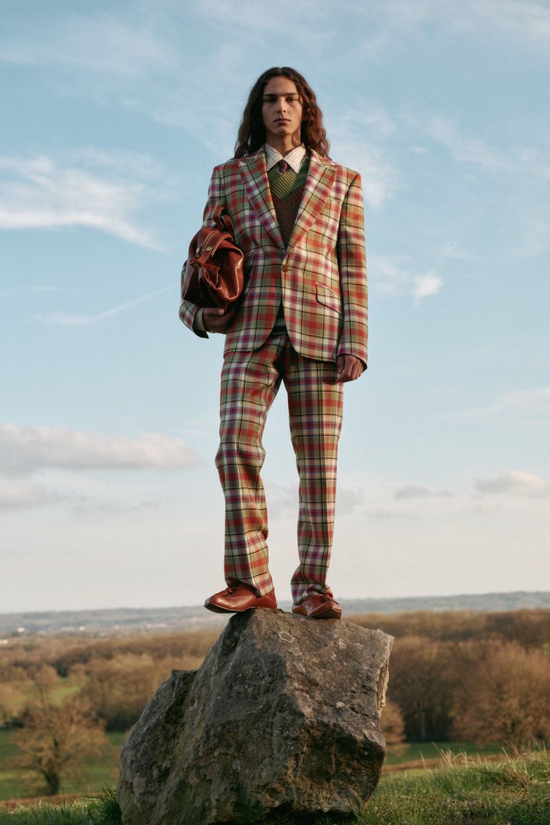 A long-haired male fashion model poses on a rock in an outdoor setting, wearing a stylish plaid suit with a leather bag.