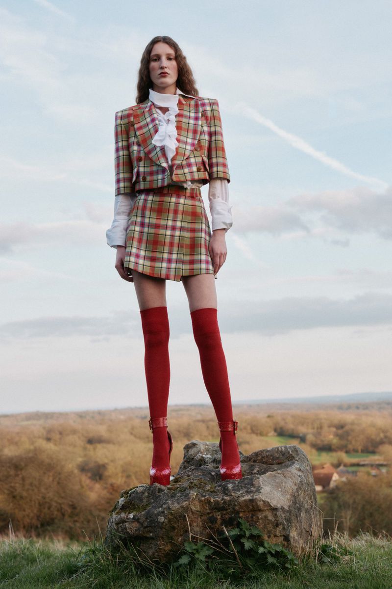 Woman in plaid skirt suit with red thigh-high boots on a rock in a field, against a cloudy sky, fashion editorial outdoor shot.