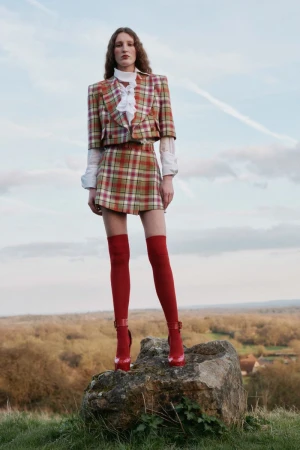 Woman in plaid skirt suit with red thigh-high boots on a rock in a field, against a cloudy sky, fashion editorial outdoor shot.