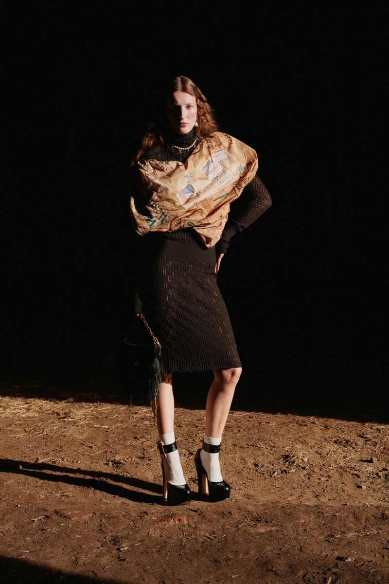 Fashion photo of a woman in a brown top, black skirt, white socks, and black platform heels posing against a dark background.