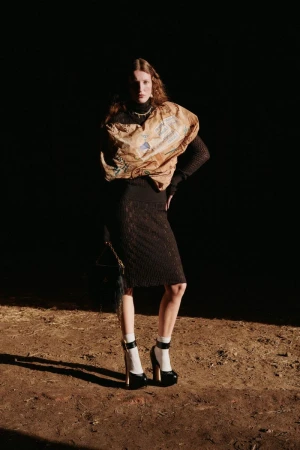 Fashion photo of a woman in a brown top, black skirt, white socks, and black platform heels posing against a dark background.