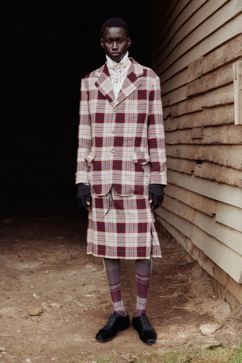 Fashionable adult male in a plaid suit and kilt stands outdoors against a weathered wooden building, showcasing editorial style.