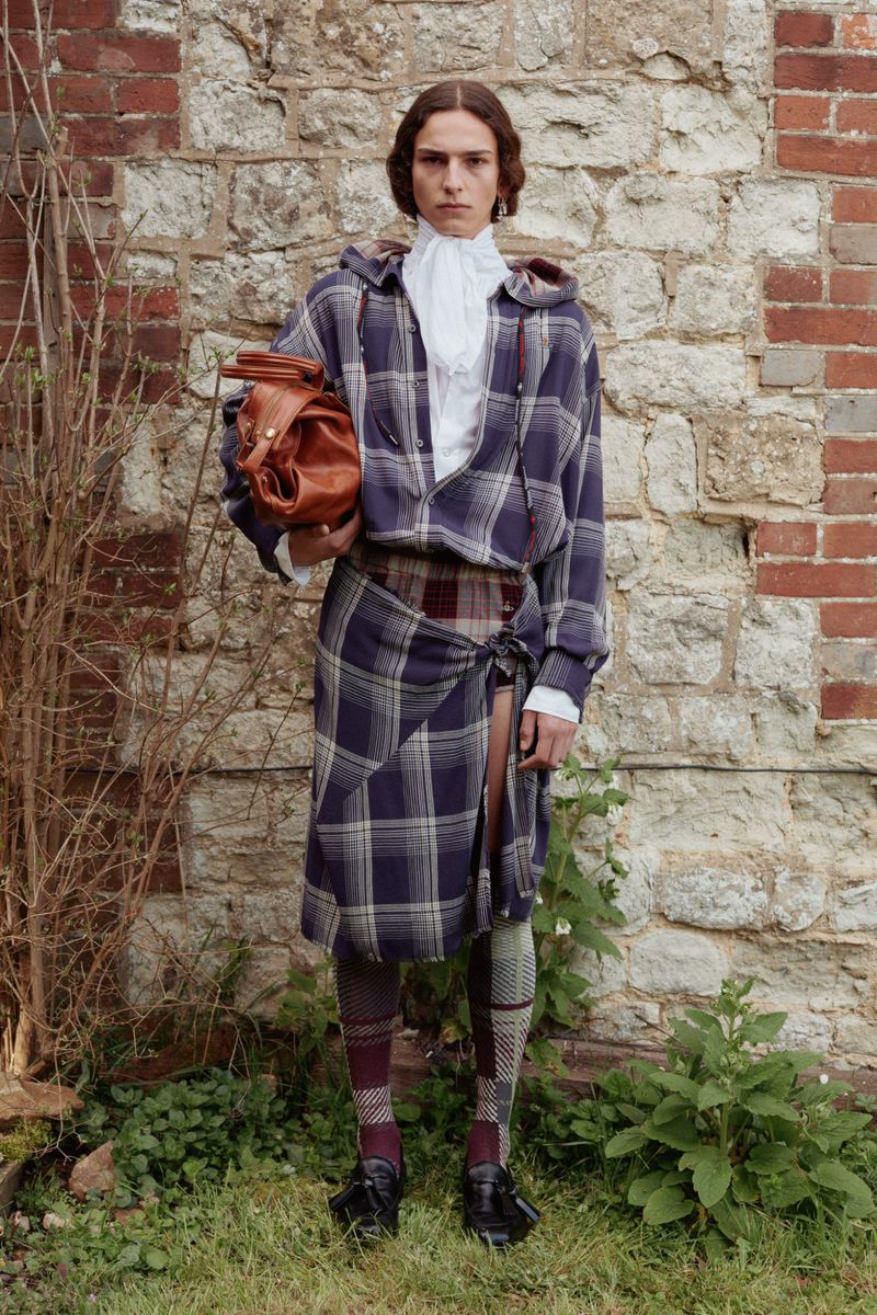 Male model poses outdoors wearing a plaid hooded jacket, kilt, patterned socks, and black shoes, holding a brown leather bag.