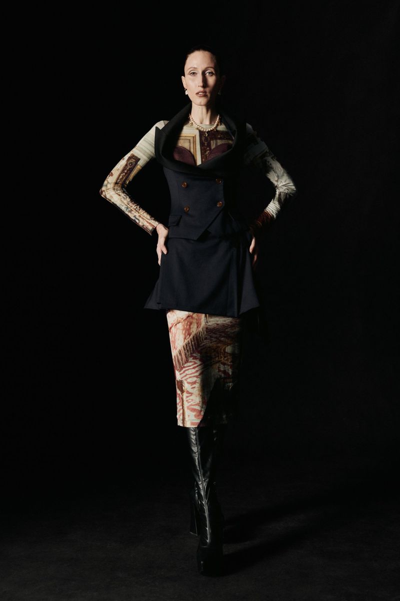 A slender adult woman poses in a sleeveless blazer, patterned top, and high-heeled boots against a dark background in a studio shot.