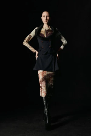 A slender adult woman poses in a sleeveless blazer, patterned top, and high-heeled boots against a dark background in a studio shot.