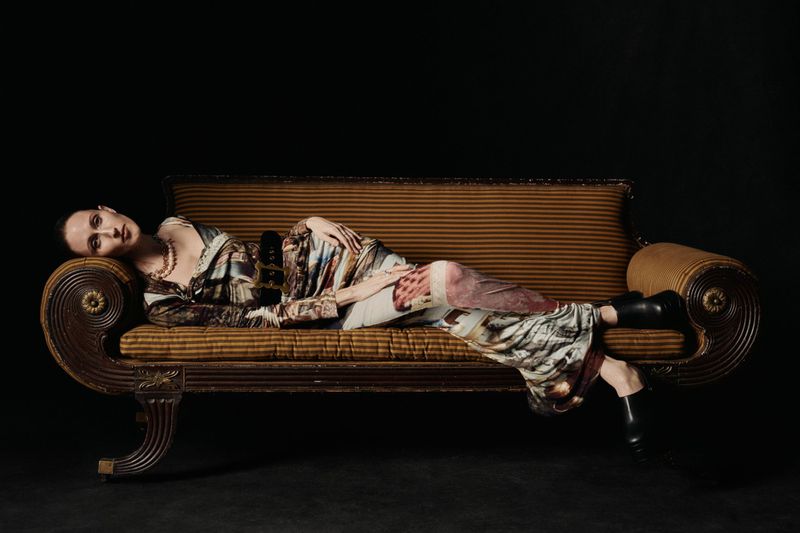 Adult female laying on a vintage-style couch with brown striped upholstery against a dark backdrop in a studio setting.