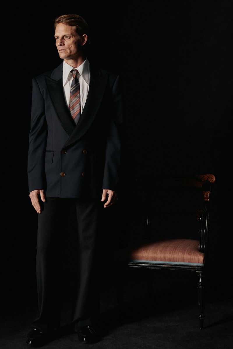 Portrait of a man in a suit looking off to the side, standing next to a chair in a dark studio with dramatic lighting.