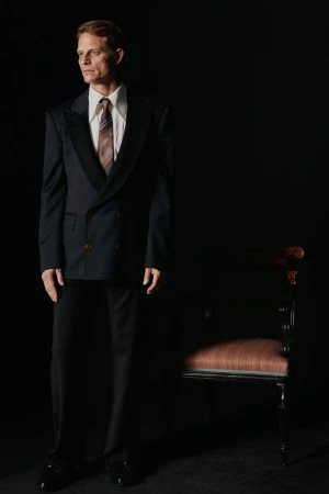 Portrait of a man in a suit looking off to the side, standing next to a chair in a dark studio with dramatic lighting.