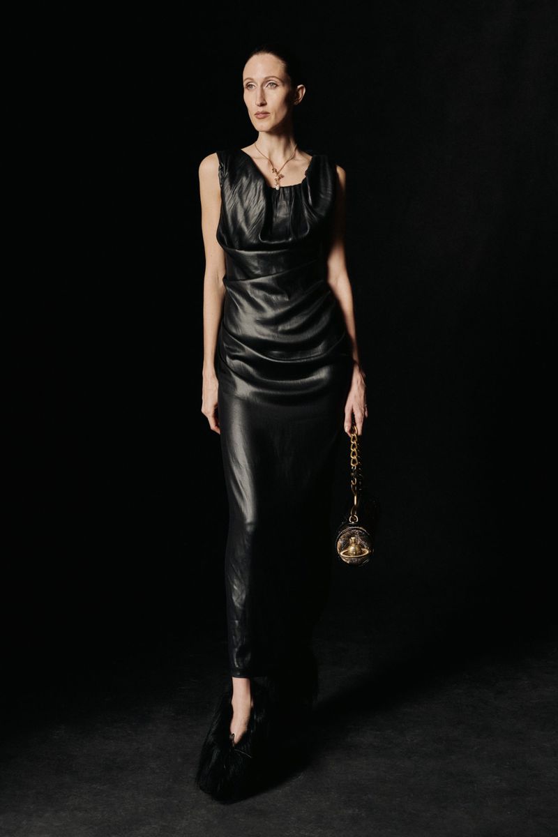 Full-length studio shot of a slim adult woman in a black sleeveless gown with a gold chain purse against a dark backdrop.