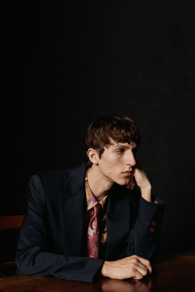 Portrait of a young adult man wearing a blazer, shirt, and tie while seated at a wooden table against a dark background.