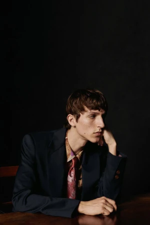 Portrait of a young adult man wearing a blazer, shirt, and tie while seated at a wooden table against a dark background.