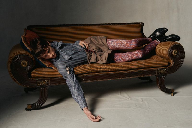 Person lying on a chaise lounge, wearing striped shirt, patterned tights and black boots, with an arm extending towards the floor.