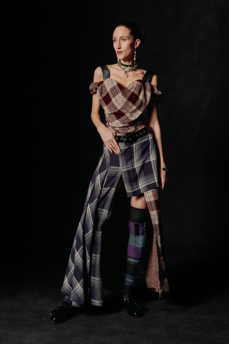 Full-length portrait of a slim adult woman in plaid top and split skirt, accessorized with layered necklaces, in a studio with a black backdrop.
