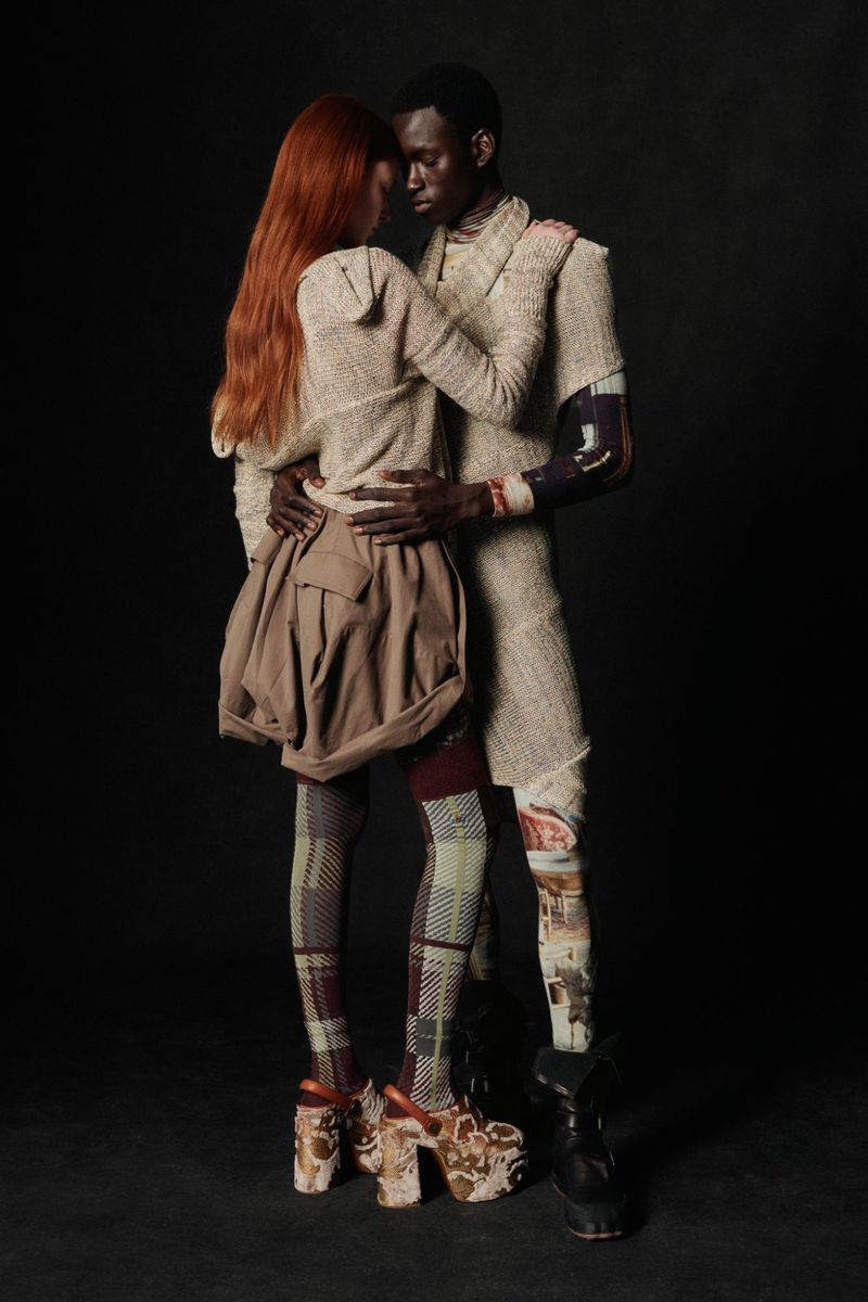 Two models in a studio embrace. One woman with red hair wears a brown skirt and patterned leggings, while a man wears textured clothing.