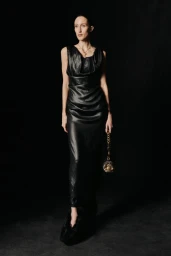 Model standing against a dark background wearing a long black leather dress and holding a small chained handbag.