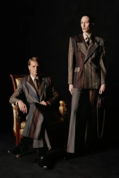 Two models wearing tailored striped suits by Vivienne Westwood, with one seated in an armchair and the other standing.