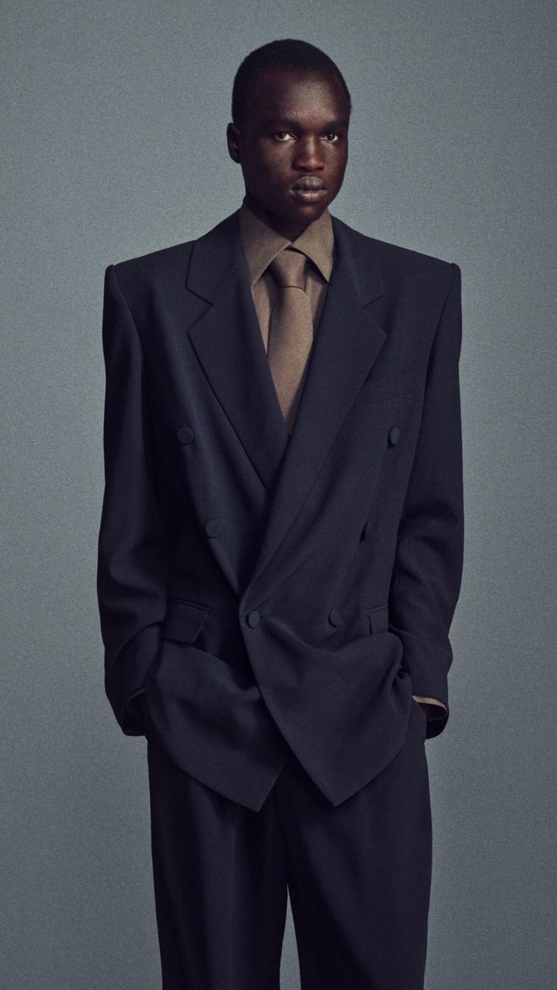Man in a dark suit with hands in pockets poses in a studio, showcasing fashion and portrait photography with classic lighting and a neutral backdrop.