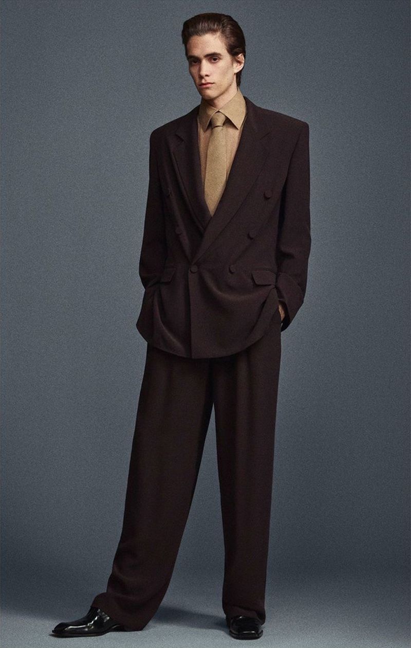 Man posing in a dark brown double-breasted suit with wide-leg pants, tan shirt and tie against a gray background in a full-body shot.