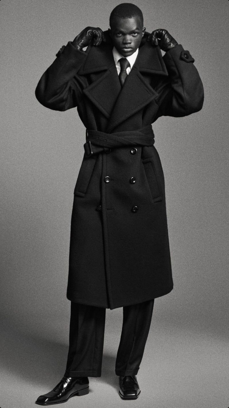 Fashionable African male model wearing a long black coat, dark suit and gloves poses in a studio with a neutral gray backdrop.