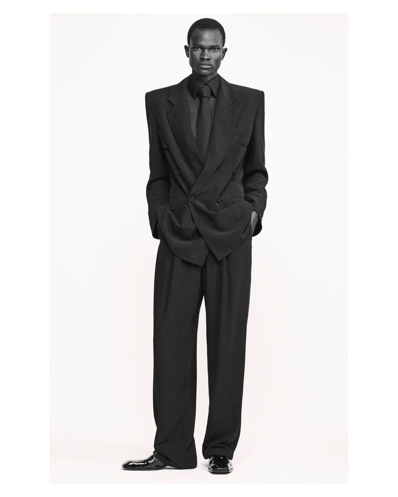 A full shot of an adult Black male fashion model in a black suit with padded shoulders against a neutral background in a photo studio.