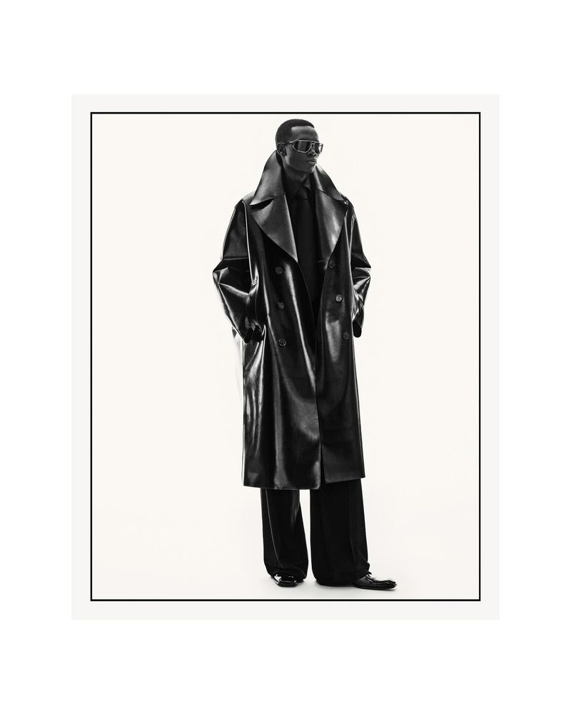 Black male model wearing an oversized black leather trench coat and dark sunglasses against a seamless white studio background.