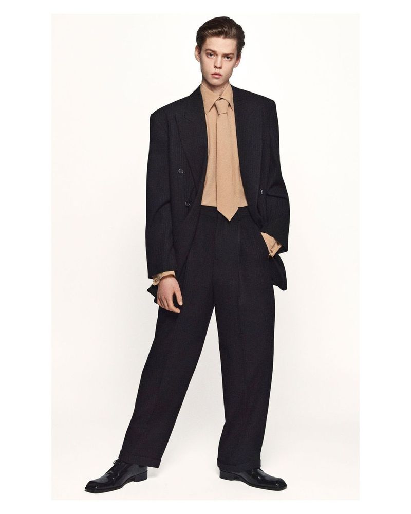 Full-length studio shot of a young adult man in a double-breasted black suit and a beige tie and dress shirt against a white backdrop.