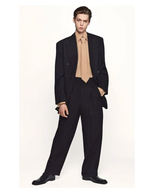 Full-length studio shot of a young adult man in a double-breasted black suit and a beige tie and dress shirt against a white backdrop.