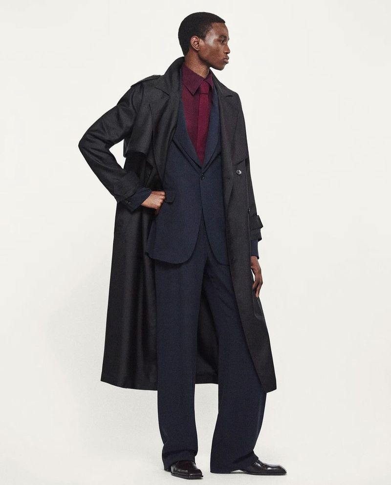 An adult Black male poses in a full-length shot, wearing a dark overcoat, navy suit, and burgundy shirt, set against a neutral backdrop.